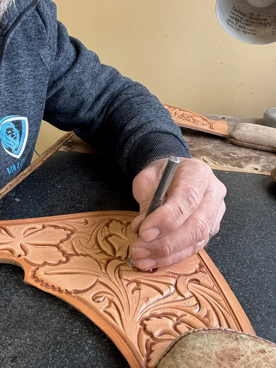 Hands tooling a floral pattern into a saddle part at the workbench