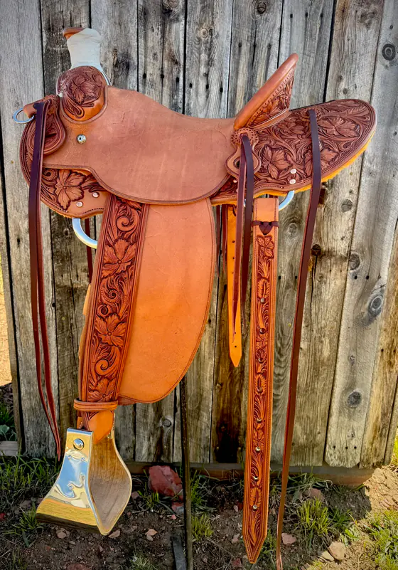 Custom Van Dyke saddle with hand-carved floral tooling on the corners and fenders, standing against a weathered barn wall