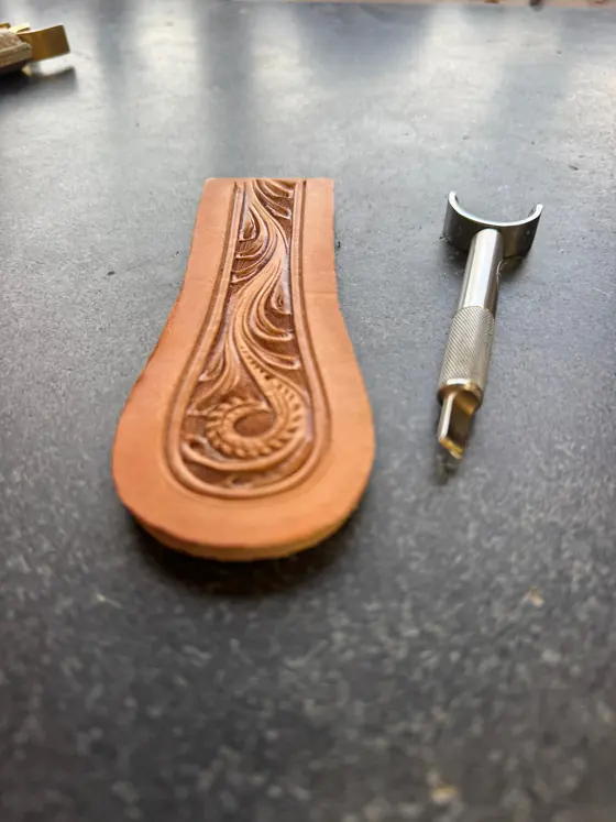 A small tooled leather piece beside a swivel knife on the workbench