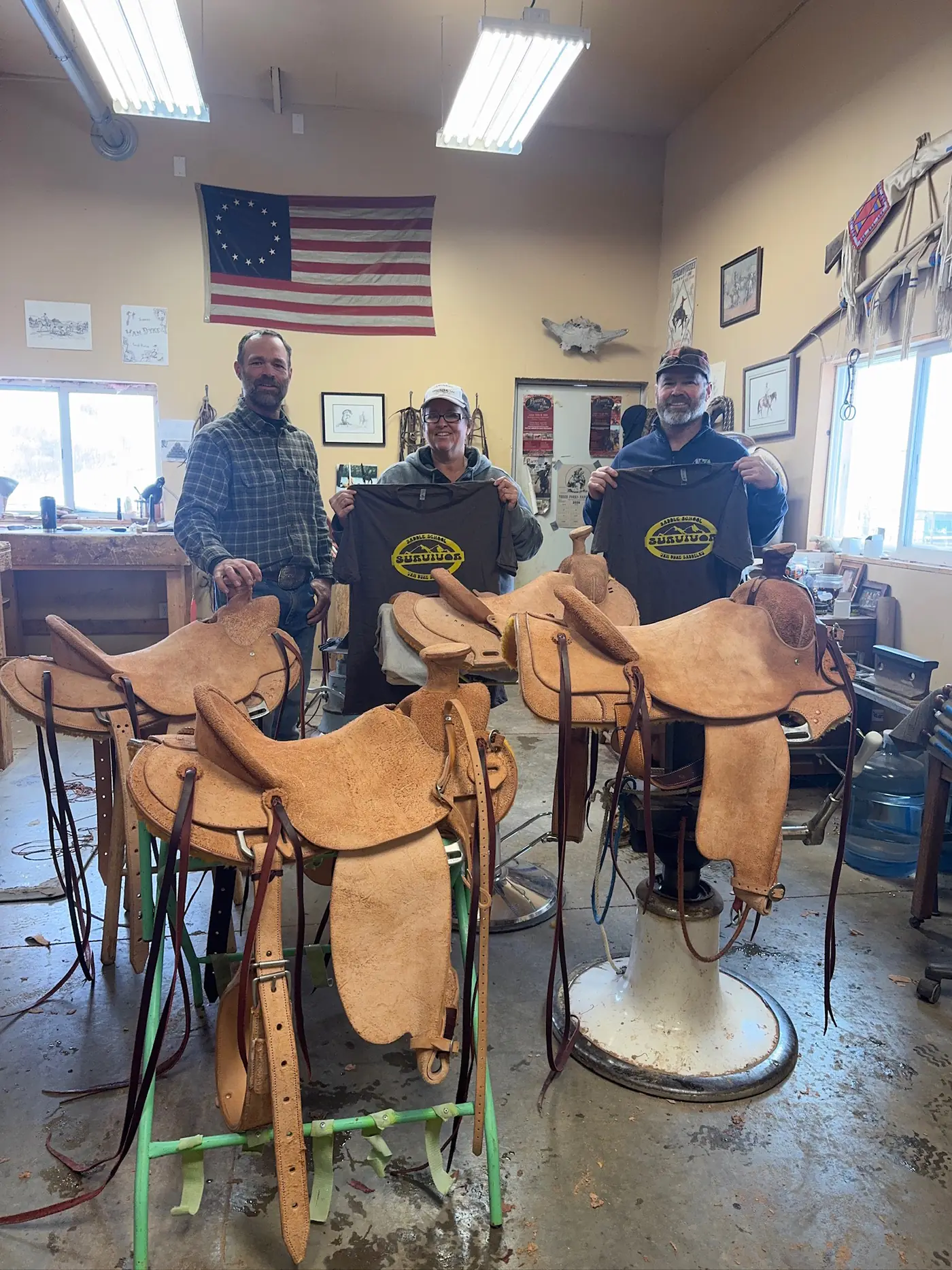 Sheridan School of Saddlery graduates holding shop shirts beside the saddles they built